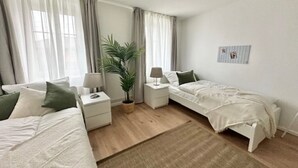 A modern bedroom with two beds, a nightstand, a lamp, a potted plant, and a small rug.