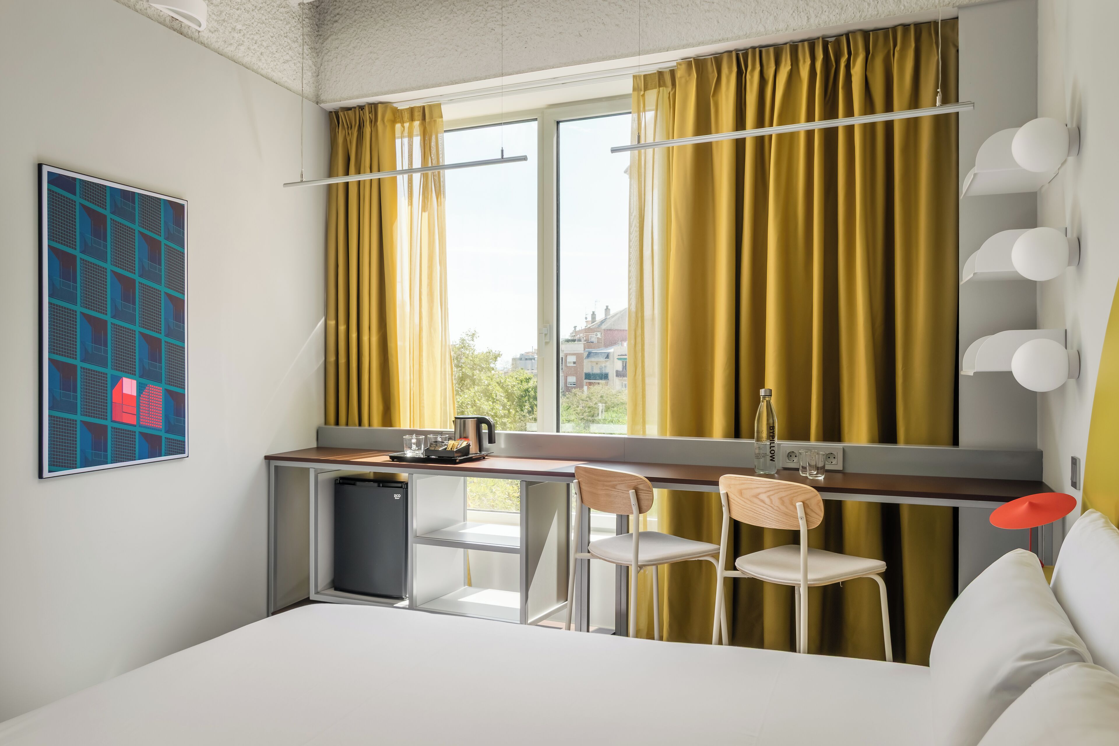 A modern hotel room with a bed, a small kitchenette, a desk with chairs, and a large window with curtains.