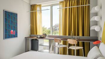 A modern hotel room with a bed, a small kitchenette, a desk with chairs, and a large window with curtains.