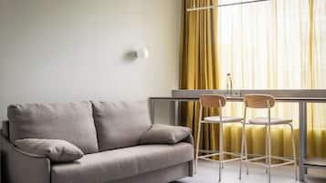 A modern living room with a grey sofa, two wooden chairs, a small table, and a window with yellow curtains.