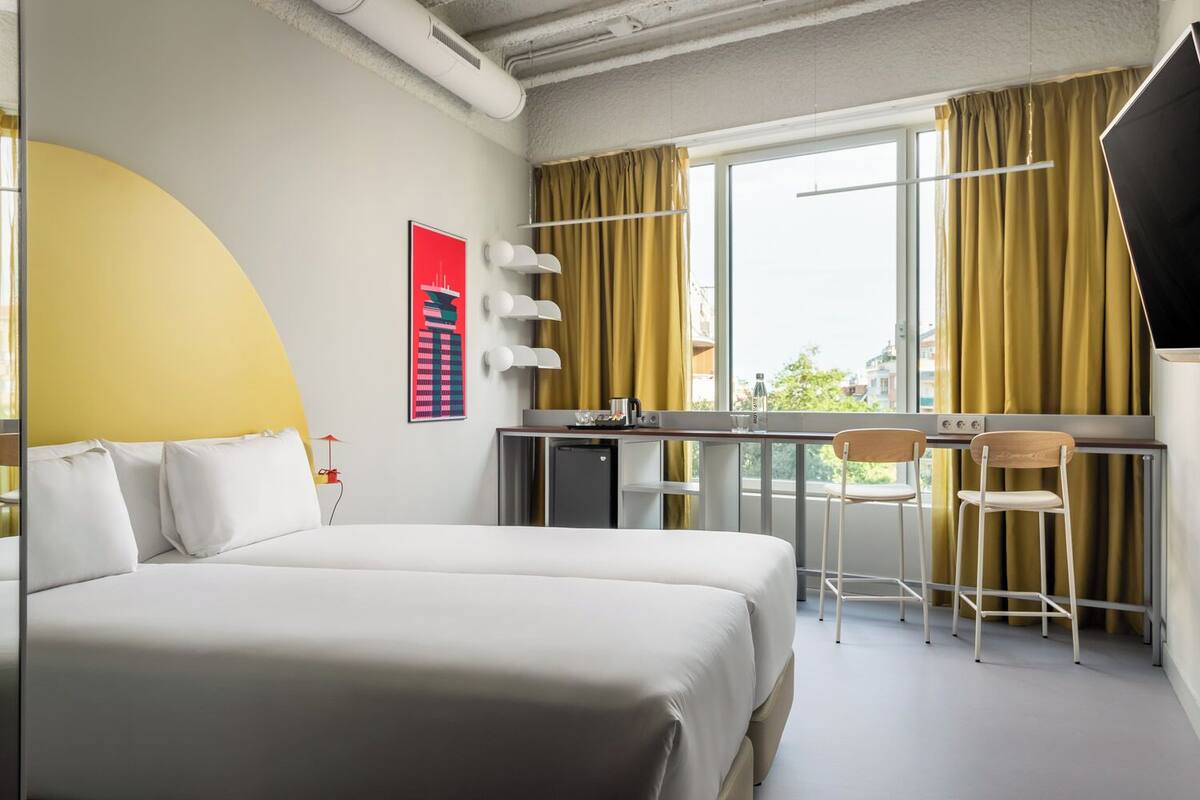 Une chambre d’hôtel avec un grand lit, un bureau avec deux chaises, une petite kitchenette et une grande fenêtre avec des rideaux.