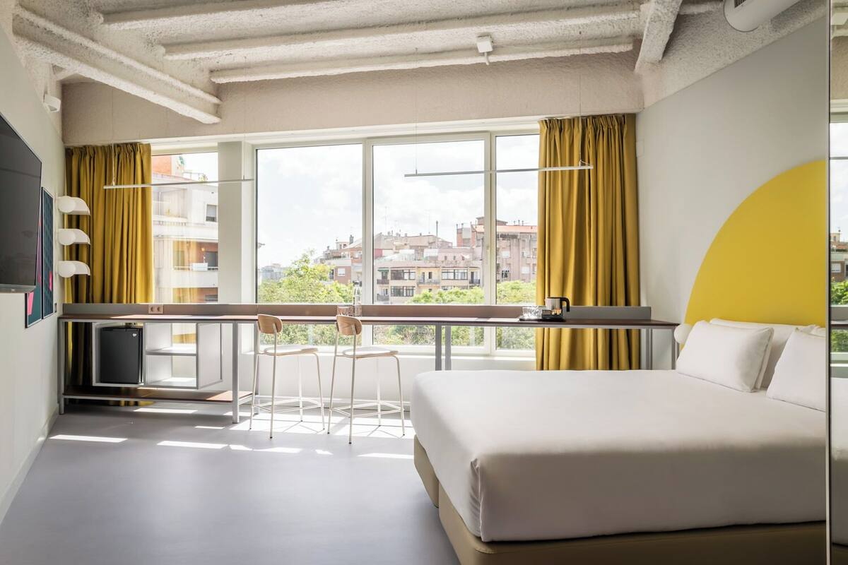 Une chambre d’hôtel moderne équipée d’un lit, d’un bureau et d’une petite kitchenette. Une grande fenêtre offre une vue sur la ville.