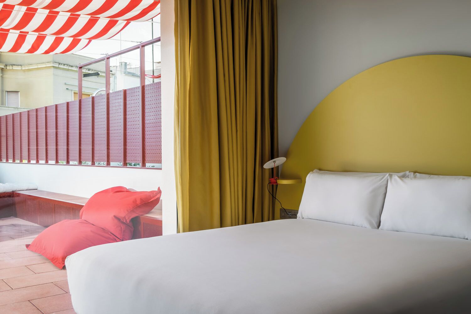 Une chambre d’hôtel avec un grand lit, des rideaux jaunes, un auvent rouge et blanc à rayures, et une vue sur un bâtiment à l’extérieur.