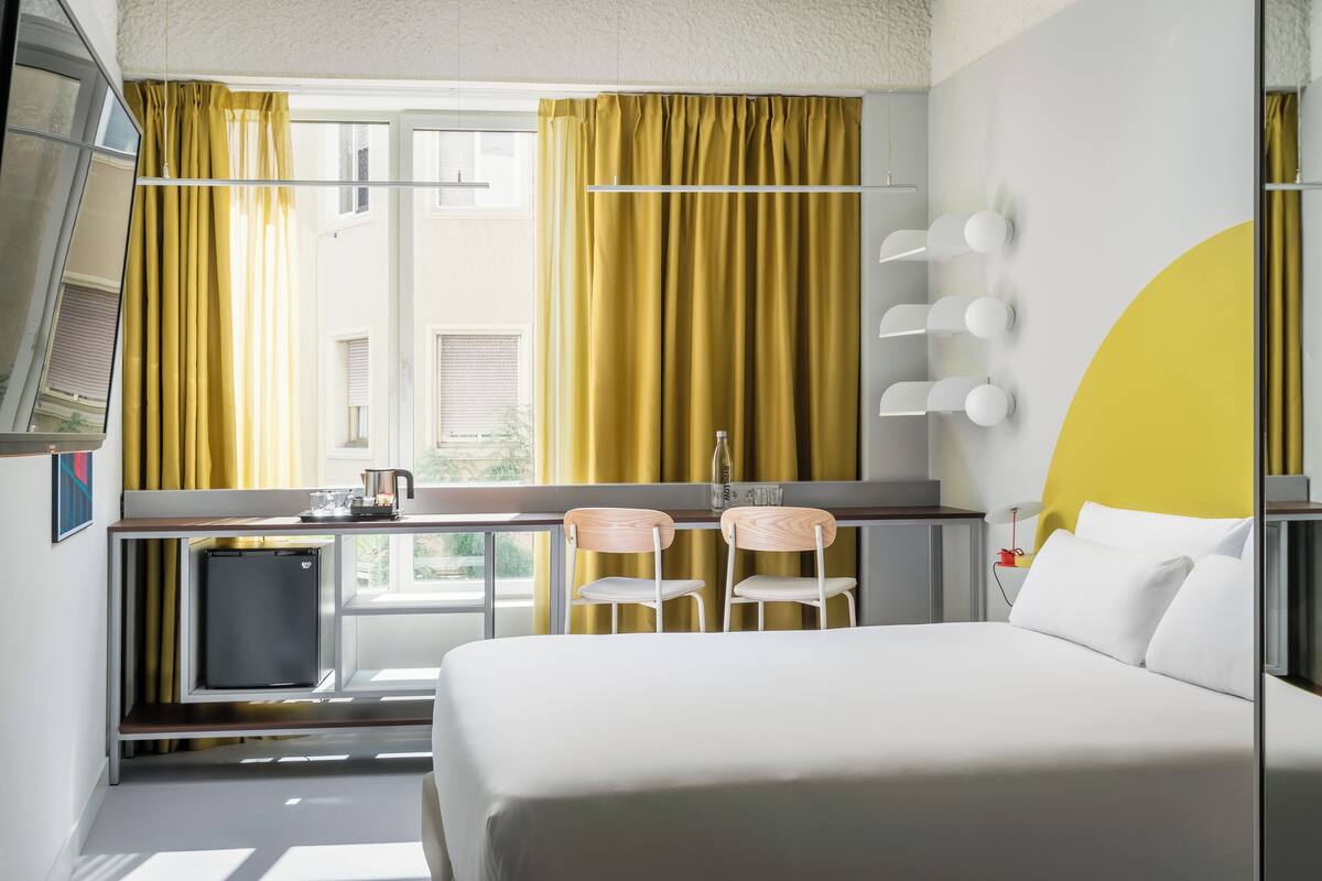 Une chambre d’hôtel moderne équipée d’un lit, d’un bureau avec un ordinateur, de deux chaises, d’un petit réfrigérateur et d’une fenêtre avec des rideaux.