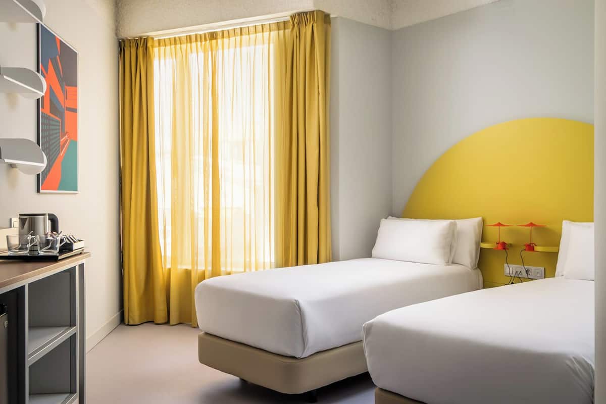 Une chambre d’hôtel avec deux lits, un bureau avec une machine à café et une grande fenêtre avec des rideaux jaunes.