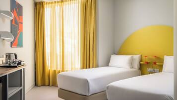 A hotel room with two beds, a desk with a coffee maker, and a large window with yellow curtains.