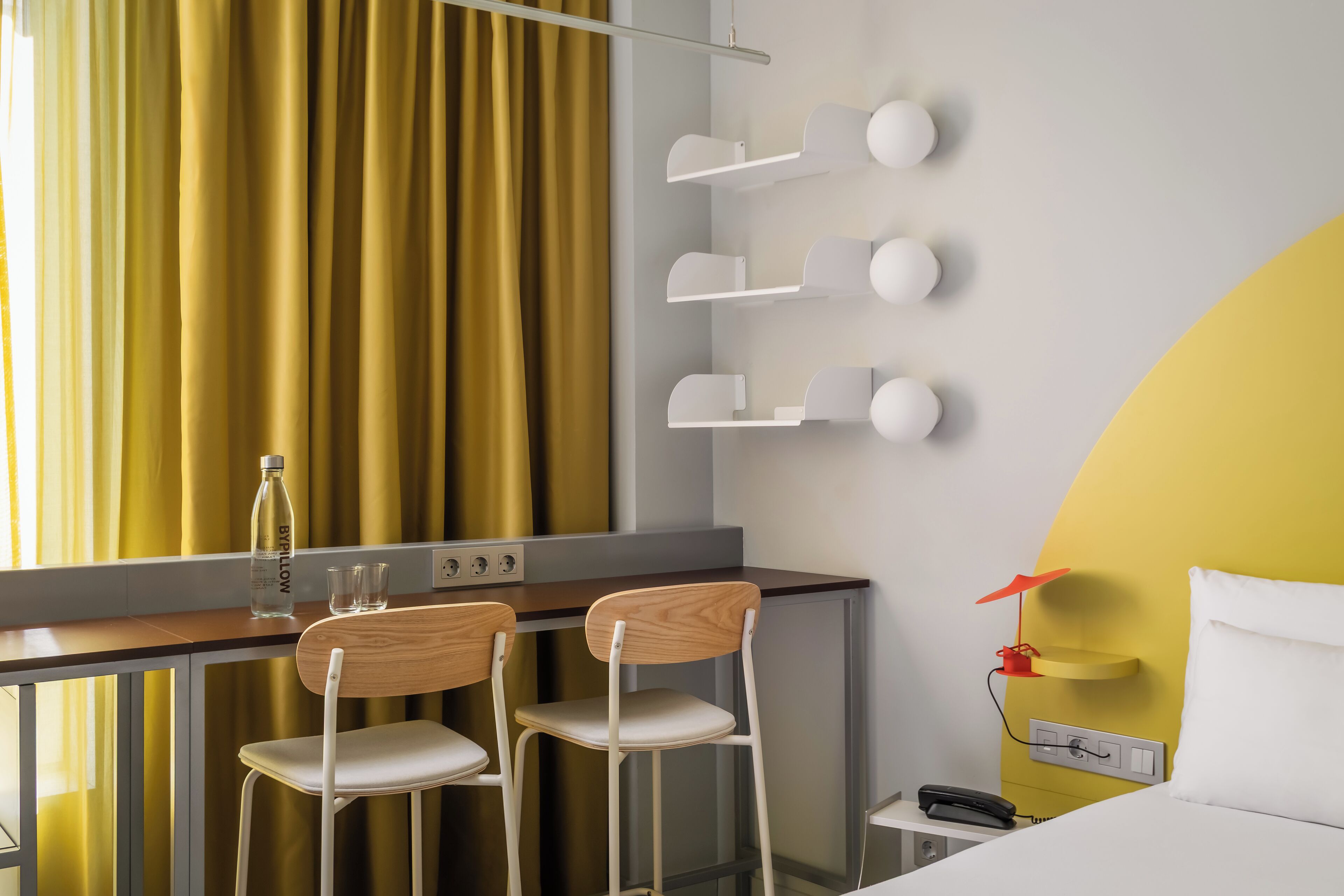 A hotel room with a desk, two chairs, a bed, a nightstand, a phone, a lamp, and a wall-mounted shelf with decorative items.