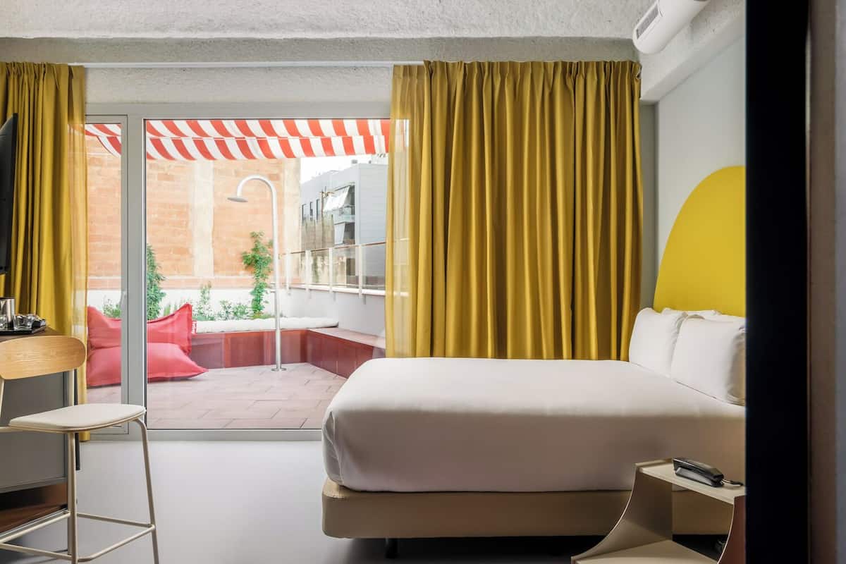 Une chambre d’hôtel moderne dotée d’un grand lit, d’un bureau et offrant une vue sur un patio extérieur agrémenté d’un auvent à rayures rouges et blanches.