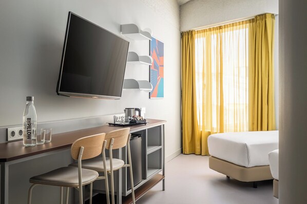 A hotel room with a TV, a desk with a bottle and glasses, a bed, and a window with yellow curtains. - BYPILLOW Flamant (Barcelona)