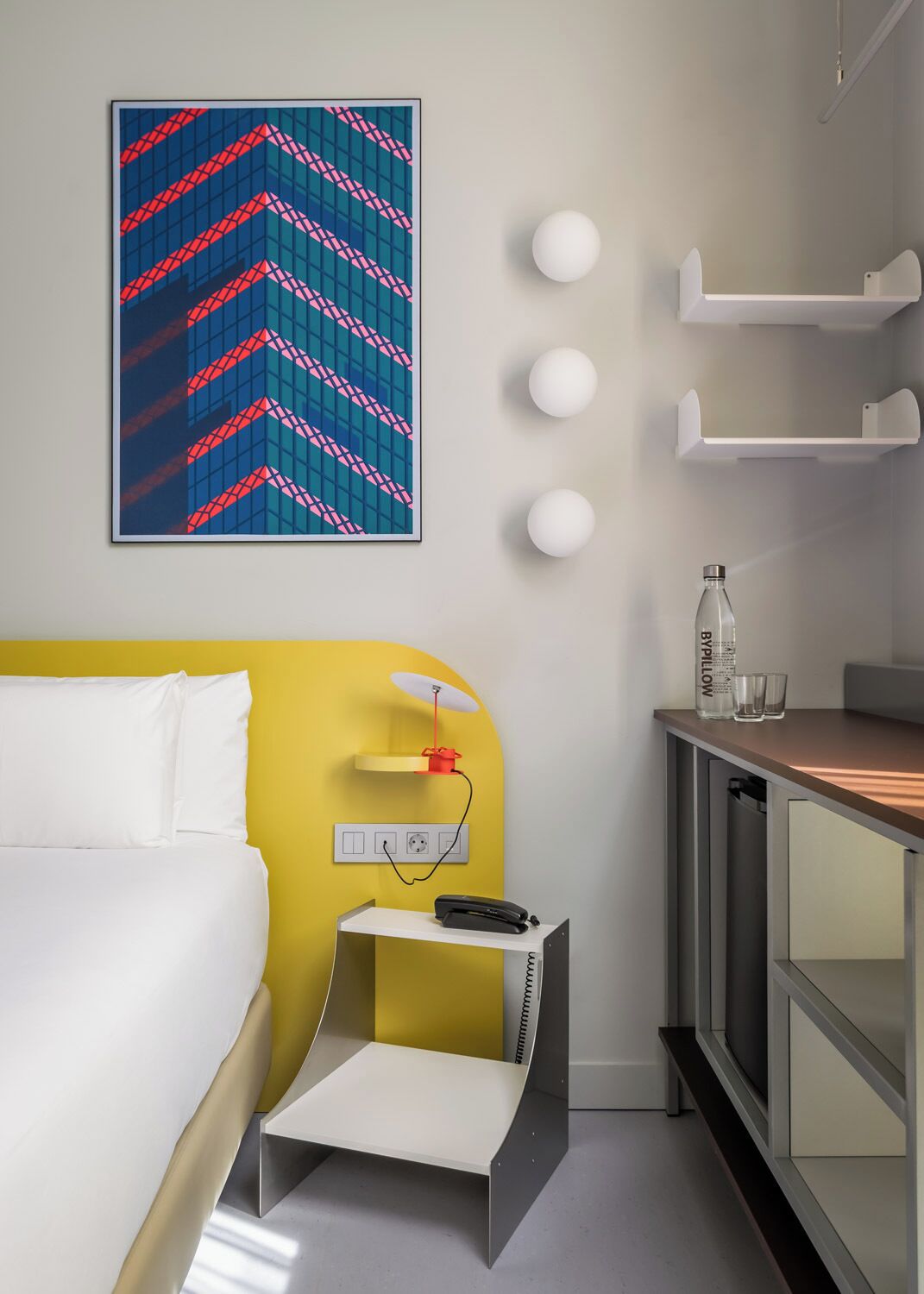 Ein Hotelzimmer mit einem Bett, einem Schreibtisch mit Stuhl, einem Telefon, einer Wasserflasche und einer Wandleuchte.