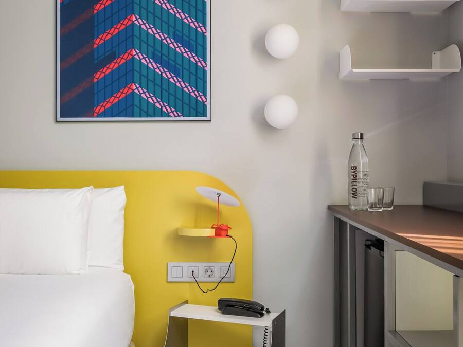 Ein Hotelzimmer mit einem Bett, einem Schreibtisch mit Stuhl, einem Telefon, einer Wasserflasche und einer Wandleuchte.