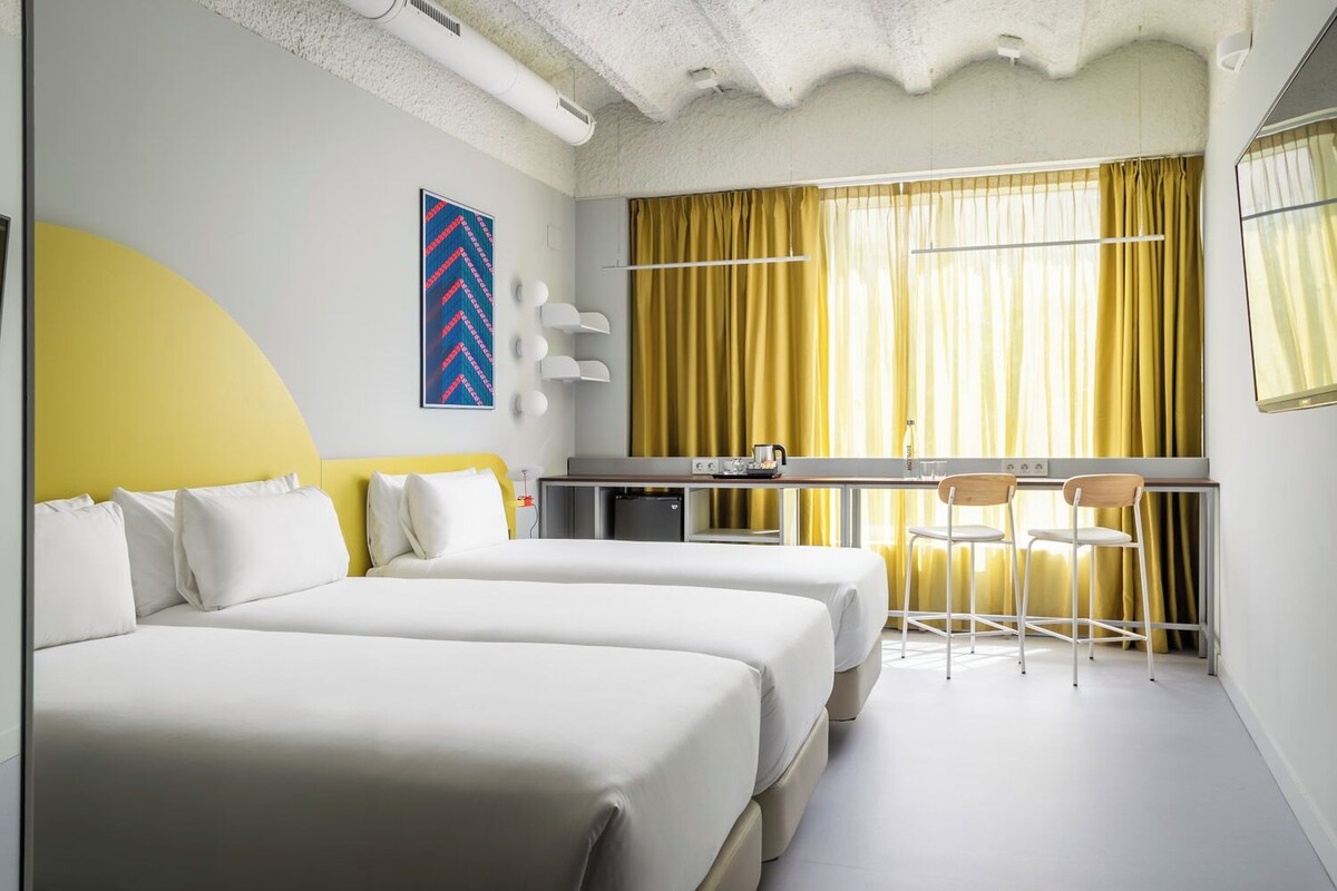 A hotel room with two beds, a desk with a chair, a large window with yellow curtains, and a modern wall art piece.
