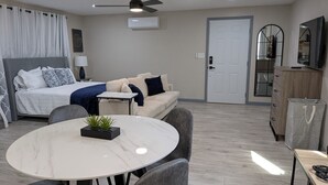 Habitació d'hotel moderna amb un llit, un sofà, una taula de dinar amb cadires, un ventilador de sostre, un aparell d'aire condicionat, un televisor i un armari.