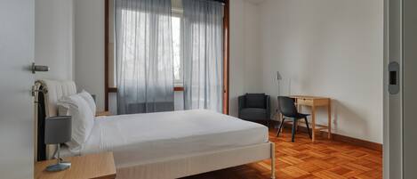 A hotel room with a bed, a desk, a chair, a lamp, and a window with curtains.