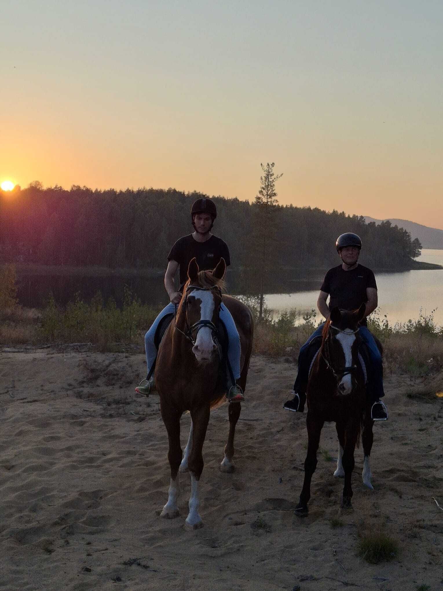 Deux personnes à cheval, évoluant sur un sentier sablonneux, avec un lac et une forêt en arrière-plan.