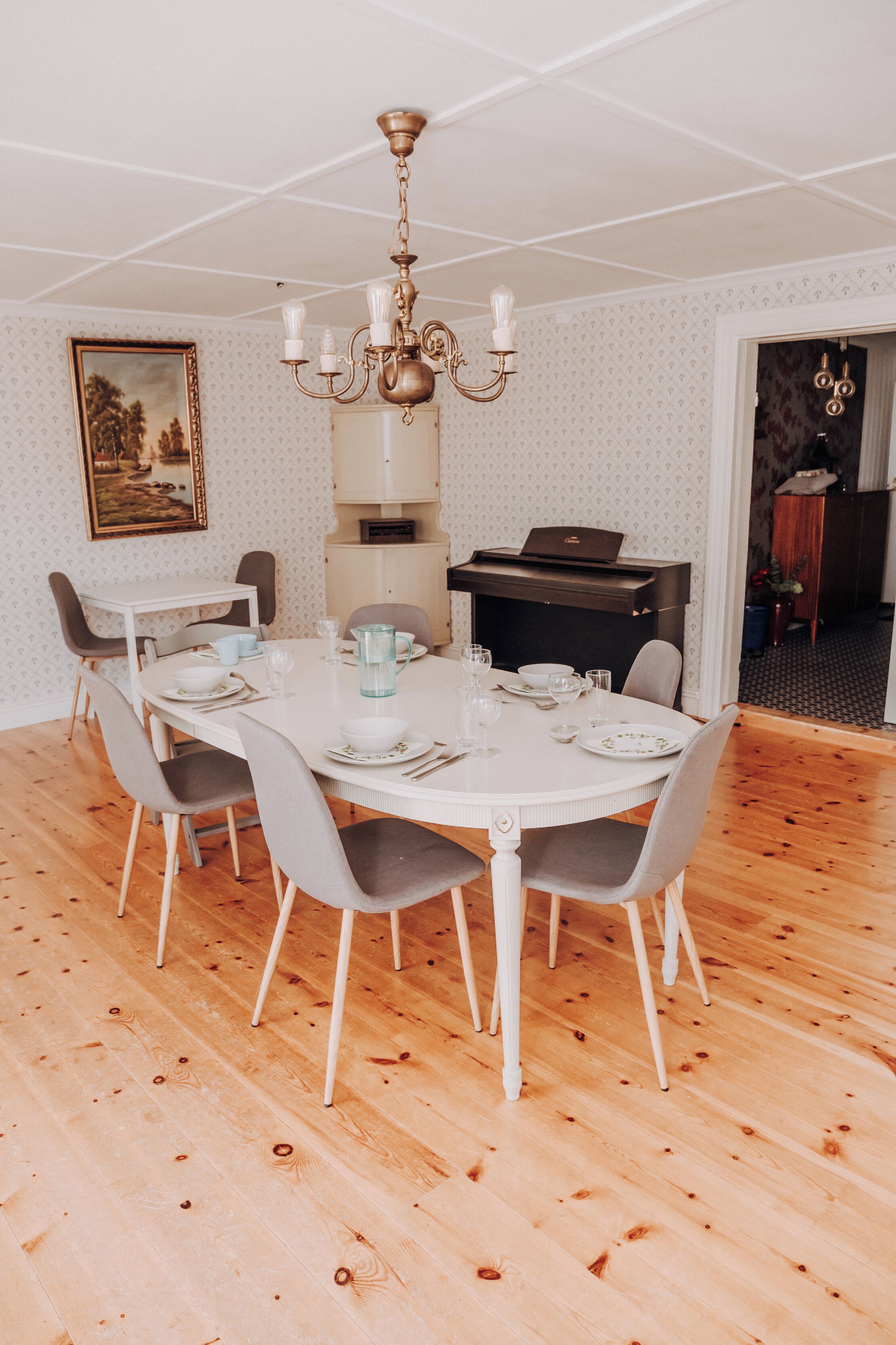 En matplats med ett runt bord, sex stolar, en kristallkrona, ett piano och en inramad tavla på väggen.