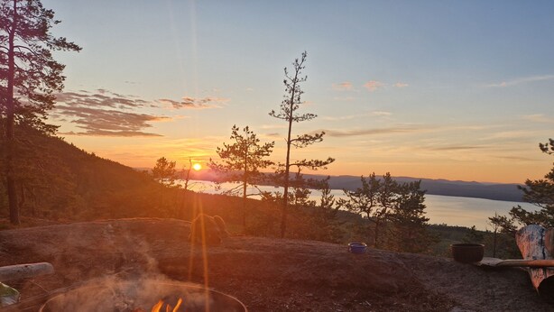 A campfire with a view of a sunset over a lake, surrounded by trees and a clear sky. - Myskje Gård Äventyr (Soderala)