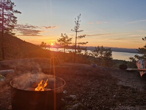 Nuotio, josta avautuu näkymä auringonlaskun värjäämälle järvelle. Nuotion ympärillä on puita ja taivas on kirkas.