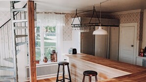 A kitchen with wooden flooring, a central island, bar stools, a hanging light fixture, and a window with a view of greenery. - Myskje Gård Äventyr (Soderala)