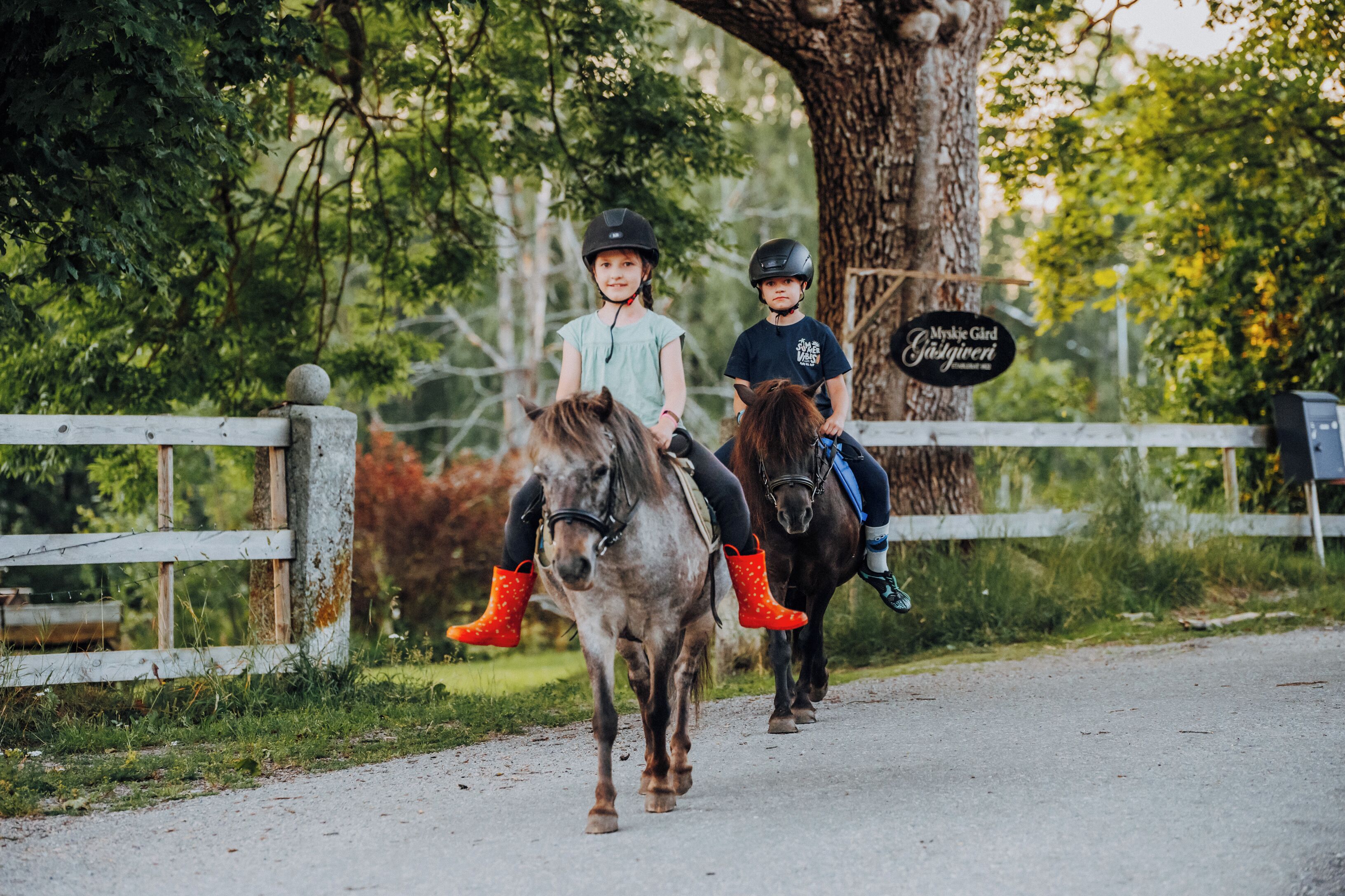 Dos niños montando a caballo por un sendero de tierra con una cerca de madera y árboles al fondo.