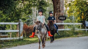 Dos niños montando a caballo por un sendero de tierra con una cerca de madera y árboles al fondo.