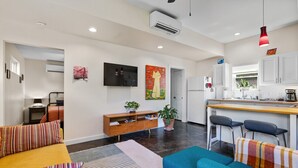 A modern living room with a sofa, a coffee table, and a TV mounted on the wall. There is a kitchen area with a counter and bar stools. - Secluded Guest House in beautiful setting (Austin)