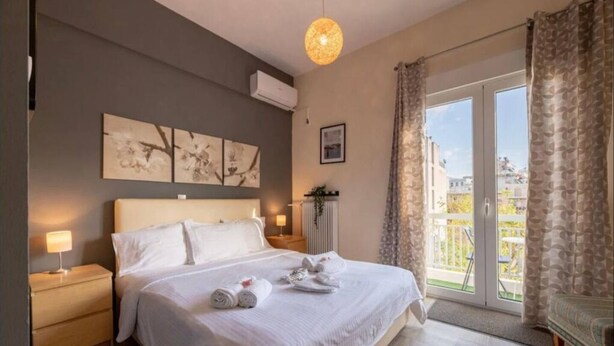 A bedroom with a large bed, a nightstand, a wall-mounted air conditioner, and a view of the outdoors through a sliding glass door. - Acropolis Vista Retreat (Athina)