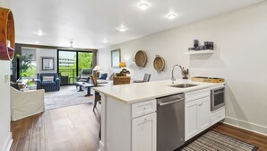 A modern kitchen with white cabinetry, a stainless steel sink, and a dishwasher. There is a dining area with a table and chairs, and a large window offering a view of the outdoors. - Sunset Rentals- 682 Mariners Way (Hilton Head Island)