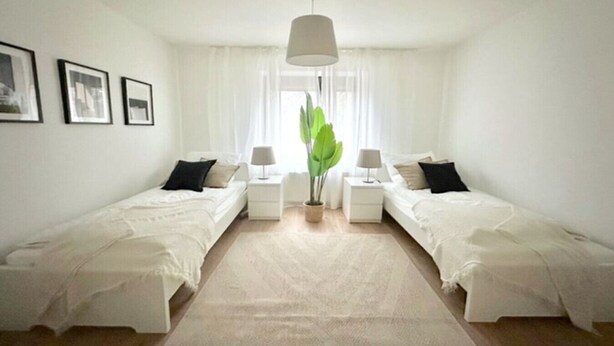 A twin bedroom with two single beds, a large window with sheer curtains, a potted plant, and a hanging light fixture.