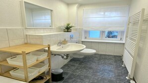A bathroom with a wooden shelf, a sink, a toilet, and a window with blinds. - Arbio Monteurzimmer Rüdersdorf 4 people (Rüdersdorf bei Berlin)
