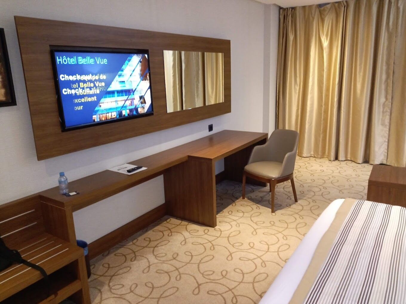Habitación de hotel con televisor de pantalla plana grande montado en la pared, escritorio de madera, sillón cómodo, cama con ropa de cama a rayas y alfombra estampada.