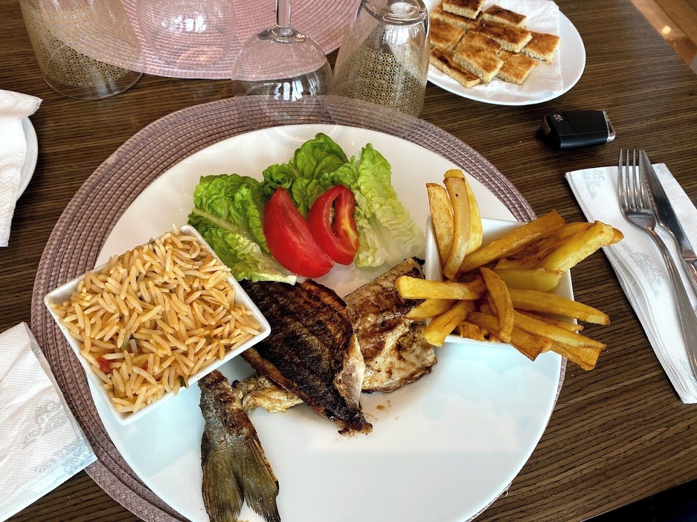 A plate with grilled fish, fries, and rice, accompanied by a salad and a sandwich.