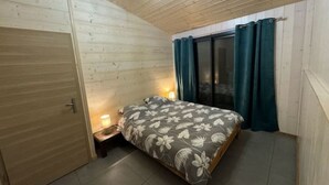 A bedroom with a wooden ceiling, a bed with a floral bedspread, a nightstand with a lamp, and a sliding glass door with curtains. - Cosy cottage with relaxing view and outdoor barbecue (Laviron)