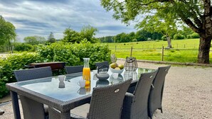 A glass-top table with wicker chairs, a pitcher of orange juice, a bowl of fruit, and a teapot. - Cosy cottage with relaxing view and outdoor barbecue (Laviron)