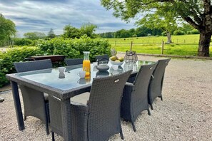 An outdoor dining area with a glass table and wicker chairs, surrounded by a gravel ground and greenery. - L'Éden – A 4-star vacation rental measuring 90 m² in the heart of a 3-hectare estate, accommodating 8 guests (Laviron)