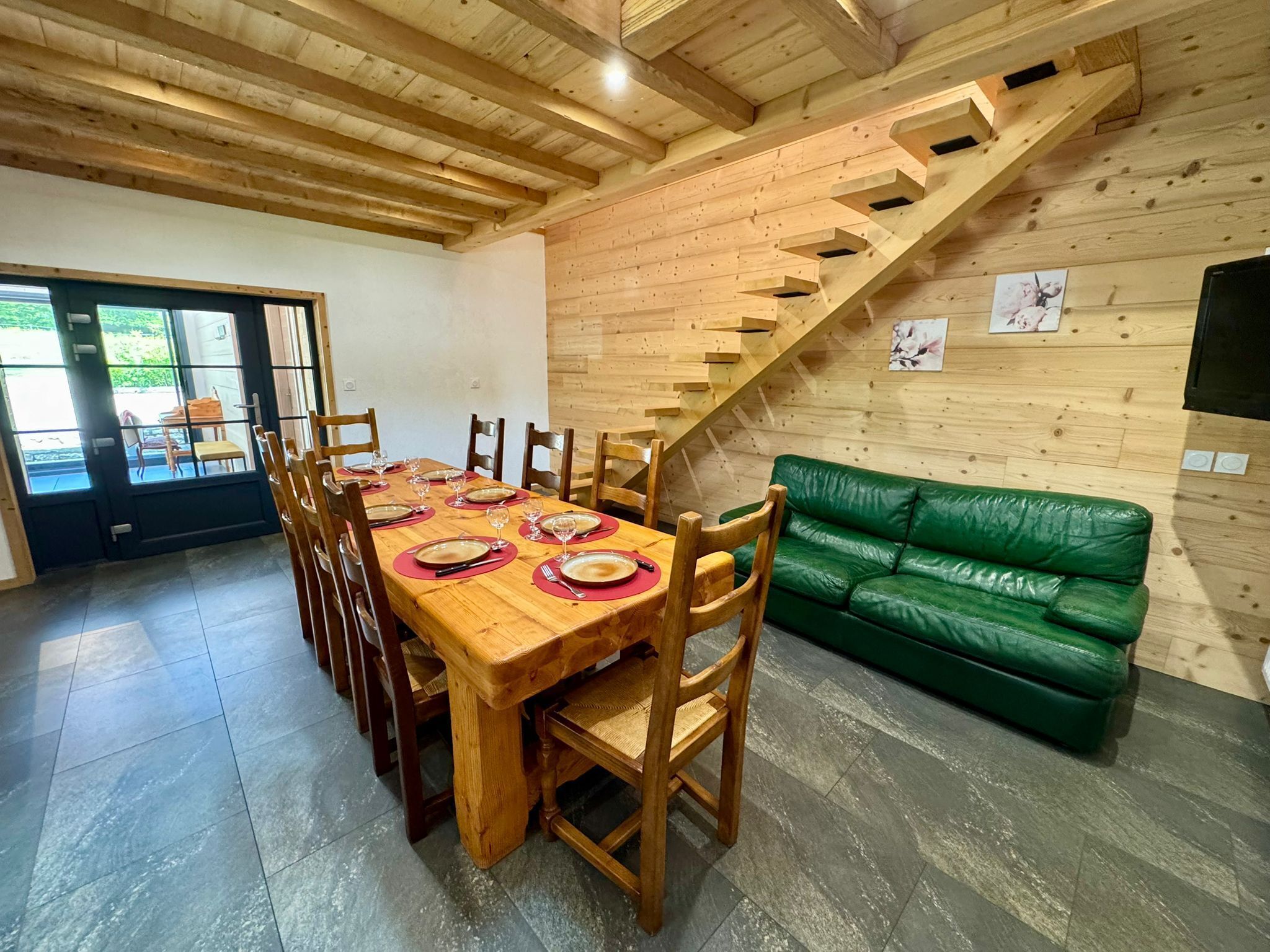 A dining area with a wooden table set for six, a green leather sofa, and a wooden staircase.