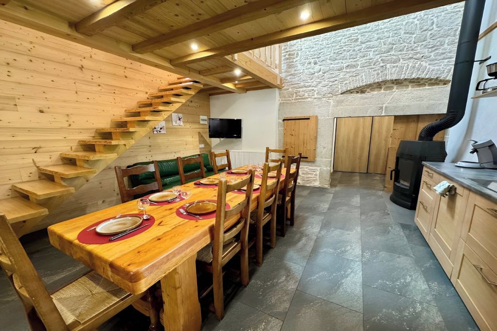 Une table à manger en bois avec des chaises, un escalier, un mur en pierre et une cheminée.