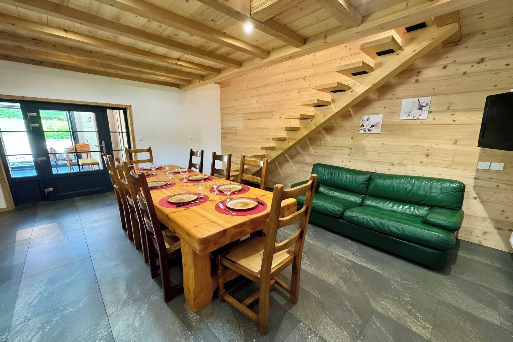Un coin repas avec une table en bois dressée pour six personnes, entourée de chaises, et un canapé vert placé contre un mur en bois, près d’un escalier.
