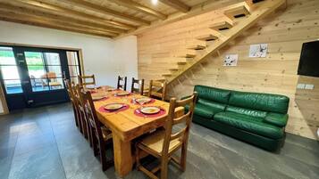 Un coin repas avec une table en bois dressée pour six personnes, entourée de chaises, et un canapé vert placé contre un mur en bois, près d’un escalier.