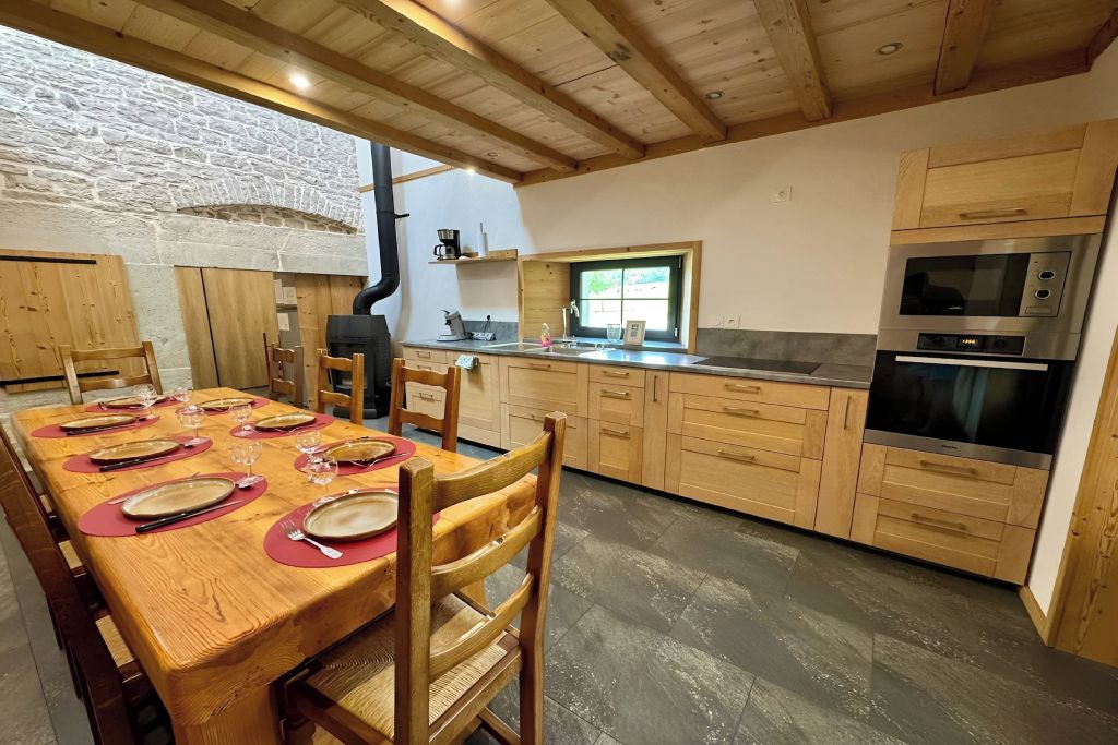 Een houten eettafel gedekt met borden en bestek, een stenen muur en een keuken met houten kasten en apparatuur.