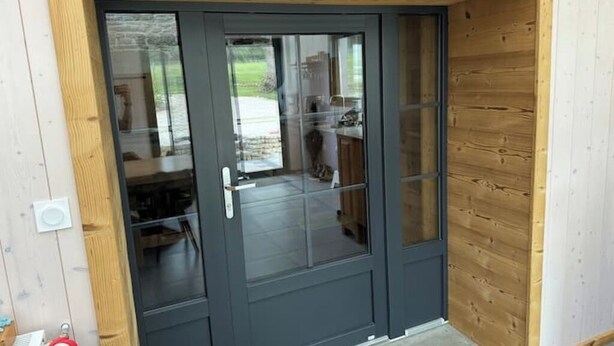A dark-colored door with glass panels, a wooden exterior, and a visible interior with a kitchen area. - Cosy cottage with relaxing view and outdoor barbecue (Laviron)