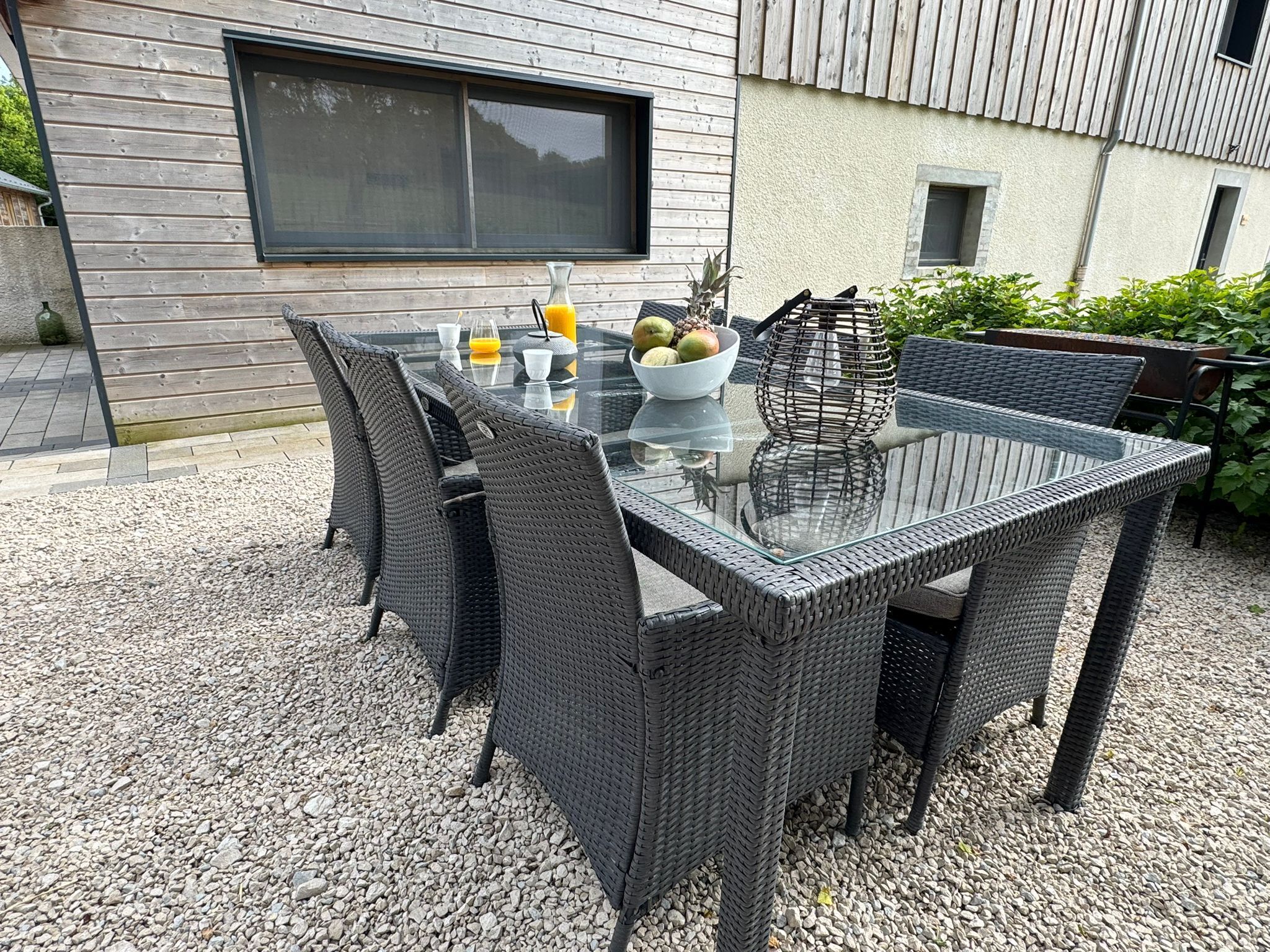An outdoor dining area with a glass-top table and wicker chairs, set on a gravel surface.