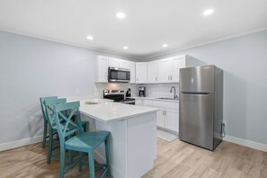 Een moderne keuken met witte kasten, een roestvrijstalen koelkast, een magnetron en een fornuis. Er staan teal-kleurige stoelen rond een klein eiland.
