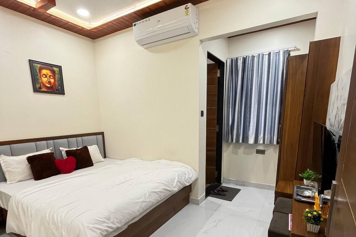 A modern bedroom with a large bed, a ceiling fan, an air conditioning unit, a small table with a plant, and a bathroom with a shower curtain.