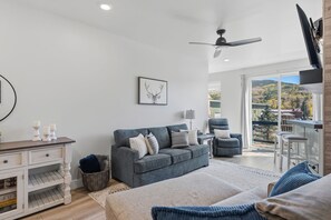A modern living room with a grey sofa, a chair, a television, and a view of a mountainous landscape through a glass door.