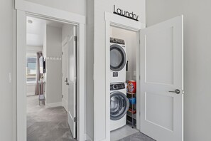 A laundry room with a washing machine and dryer, a door labeled 'Laundry,' and a glimpse of a hallway leading to a room with a window and curtains.