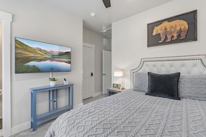 A bedroom with a large bed, a flat-screen TV mounted on the wall, a bedside table with a lamp, and a framed picture of an elephant.