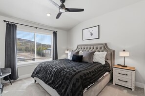 Un dormitorio con cama, ventilador de techo, mesita de noche con lámpara, un cuadro enmarcado en la pared y una ventana con cortinas.