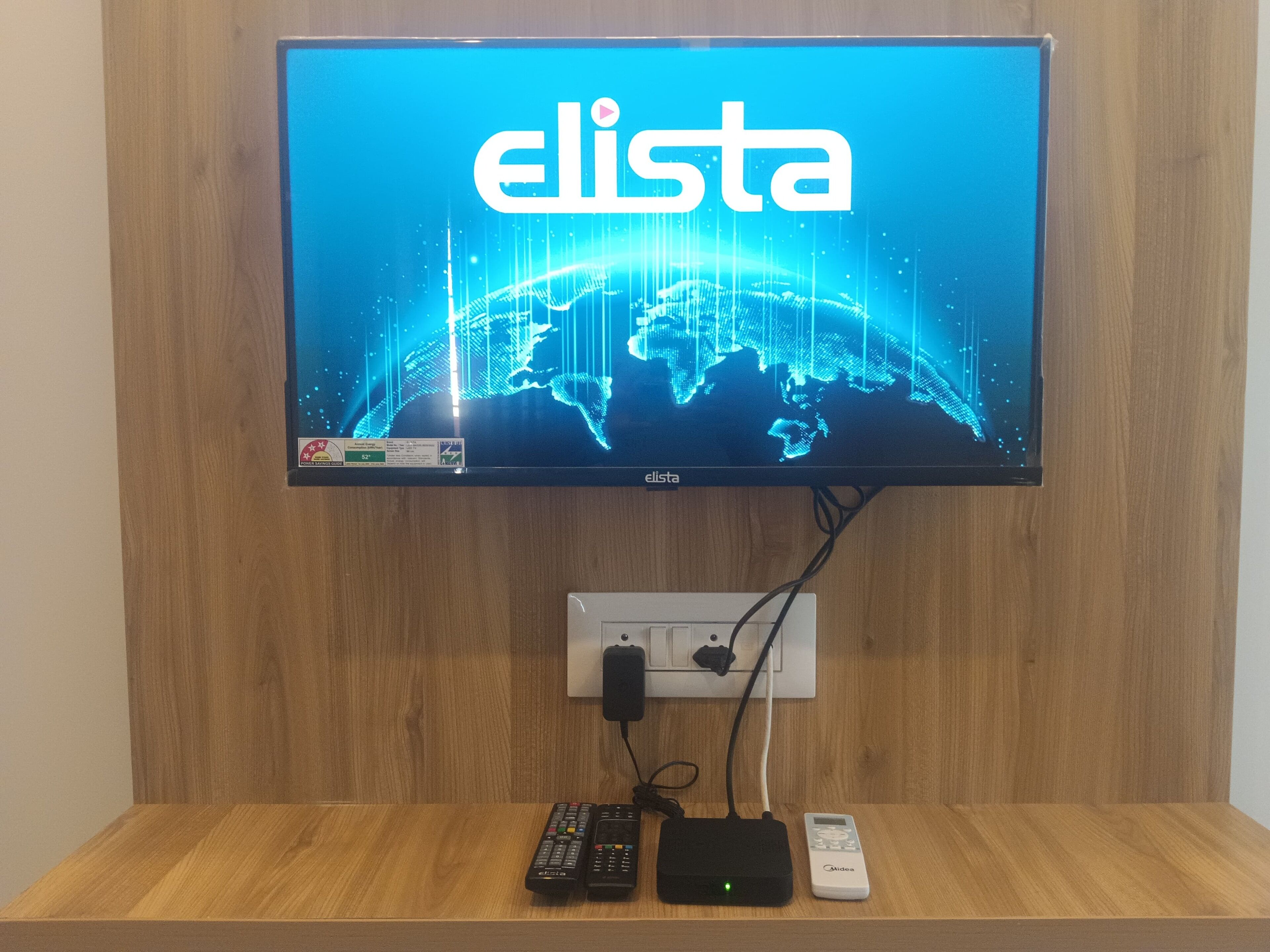 Un televisore a schermo piatto montato su un muro di legno che mostra il logo Elista. Sotto la TV, uno scaffale di legno con una ciabatta multipresa, un telecomando e un dispositivo mobile.