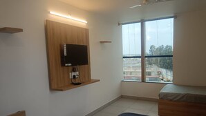 A hotel room with a bed, a TV mounted on a wooden panel, a window with a view of buildings, and a ceiling fan.